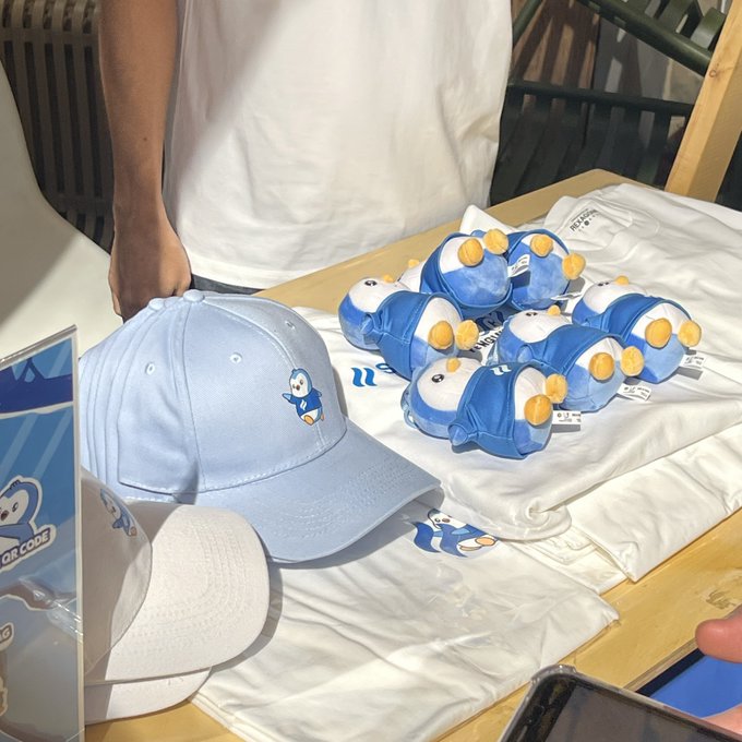 A display table features a light blue baseball cap with a penguin character logo, alongside plush penguin toys and folded white t-shirts with matching designs, with a person in a white shirt standing behind the merchandise.