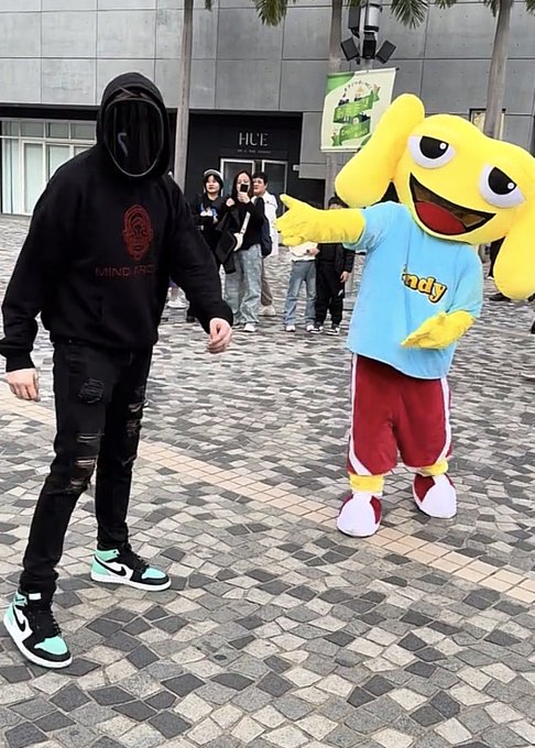 A masked figure in a black hoodie with "Mind's Eye" text stands facing a large yellow mascot wearing a "Sandy" t-shirt and red shorts, with the caption "Gm if you Gm" playing on the crypto greeting "gm" (good morning) while the two appear to greet each other in a public plaza.