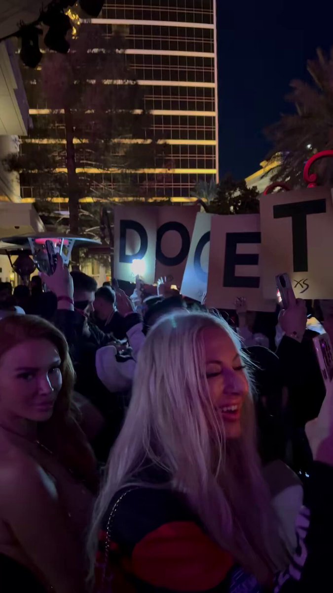The image features a woman with blonde hair wearing a red and black striped jacket, holding up a sign that has "dog" written on it. Another sign behind her reads "Doge", indicating they may be related or part of the same cause. The background shows people standing in front of a tall building, suggesting an event is taking place outside. There are also other signs among them that read "$DOGE TO ?".