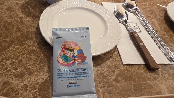 A packet of '2026 KAKASHI PHANTOM PUDGY PENGUINS TRADING CARDS' from the Pudgy Penguins NFT collection, containing 6 cards per pack, sits on a white plate beside a spoon and knife.