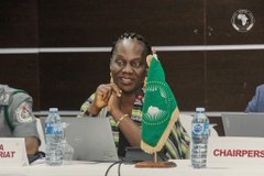 A woman wearing a "CHAI" (Chairperson) name placard smiles while seated at a conference table with a green flag, likely from the African Union or a similar regional body, suggesting a diplomatic or organizational meeting related to the #IOTA post's discussion of outcomes.