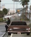A low-resolution image shows a brown classic car parked on a street with people standing in the background.