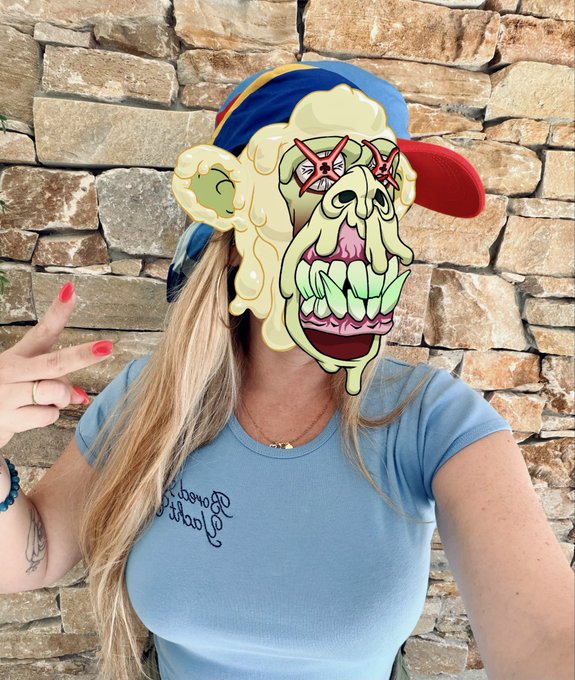A woman in a light blue 'Boerne Volleyball' t-shirt and a colorful cap poses with a peace sign, with a cartoon monster face filter covering her face against a stone wall background.