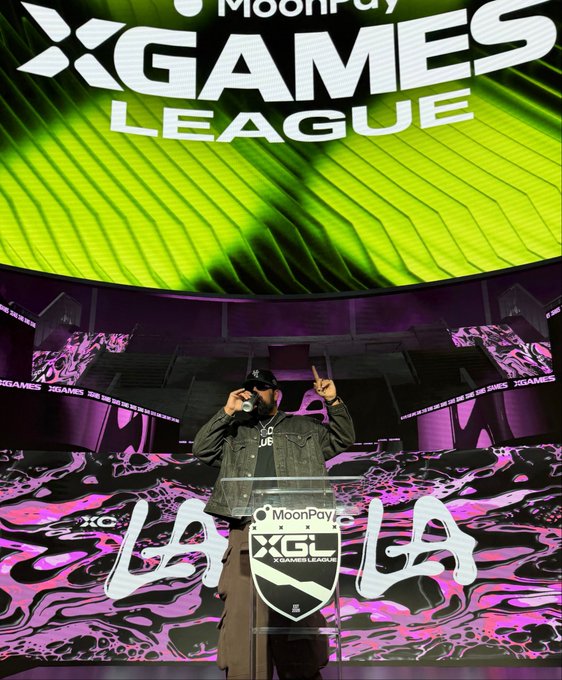 A man wearing a black cap, sunglasses, and denim jacket speaks into a microphone at a podium featuring the "MoonPay XGL X Games League" logo and "LA" text, while standing on a stage with a large "XGAMES LEAGUE" display and vibrant green and pink graphics. The image captures the speaker representing MoonPay and Solana at the X Games League event in New Orleans, aligning with the post's announcement of his appearance and request for content ideas.