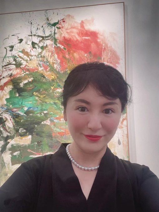 A woman with dark hair, red lipstick, and a pearl choker poses in front of a colorful abstract painting.