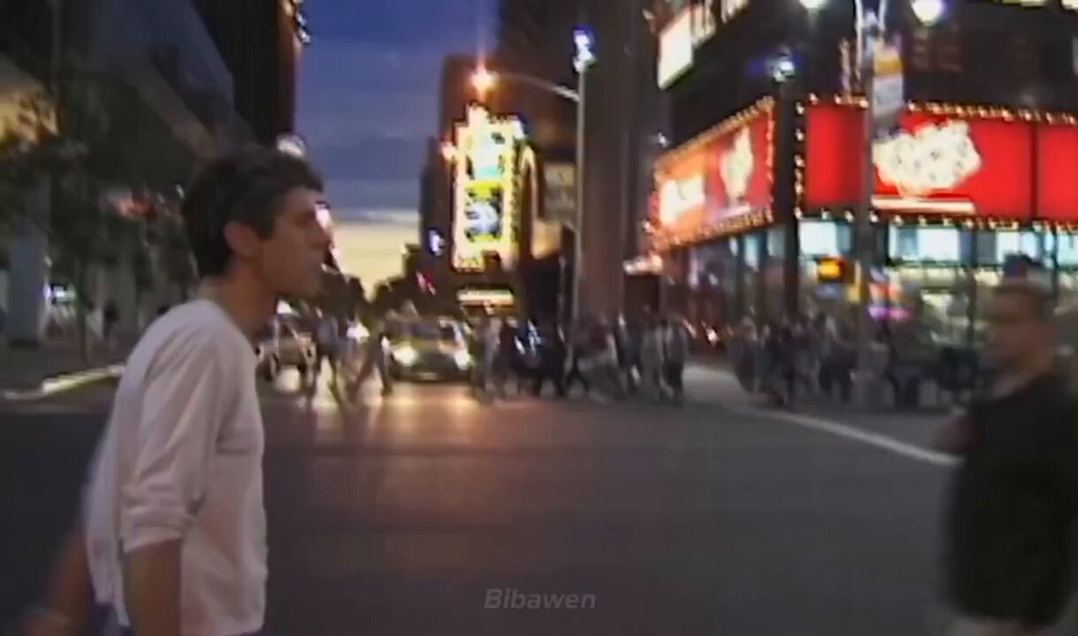 The image shows two people walking down a city street at night, surrounded by tall buildings and neon signs. The scene captures the essence of urban life with its bustling atmosphere and bright lights. The photo has been edited or filtered to emphasize certain elements while downplaying others, creating an interesting visual effect that draws attention to specific aspects of the image.