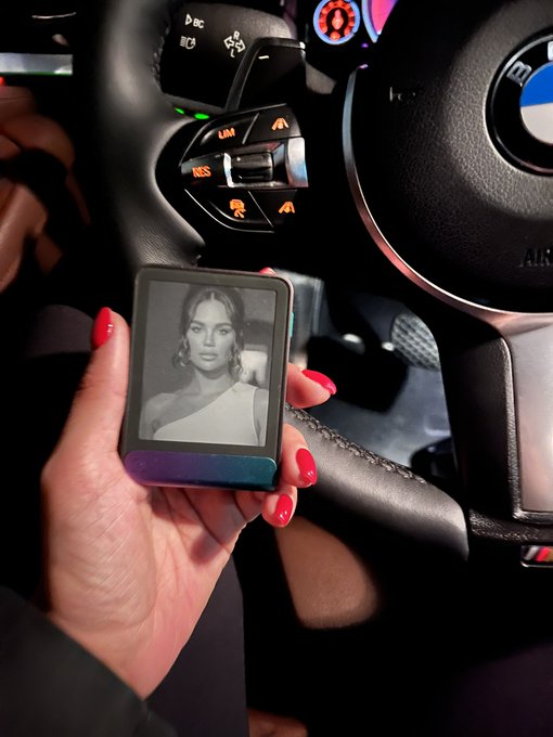 The image shows a person holding up an electronic device, possibly a smartphone or tablet, displaying a picture of a woman's face. The device appears to be black and white, which might suggest that it could be an older model. The person is wearing red nail polish on their fingers, adding a pop of color to the scene. The background features a car steering wheel, indicating that they are in a vehicle or near one. The image also contains text that reads "gm to everyone build with AI earn with crypto protect it with a Ledger stay safe out there".