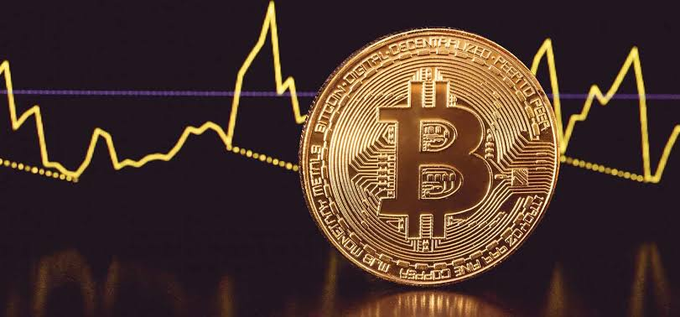 A physical Bitcoin token sits in front of a glowing yellow line chart displaying volatile price movements, with the "B" symbol clearly visible on the coin. The image visually represents the launch of Bitcoin VIX (BVIV) perpetual contracts on Hyperliquid, symbolizing the trading of Bitcoin's implied volatility.