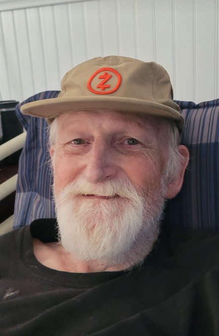 A smiling older man with a white beard wears a tan cap featuring a red circular "Z" logo, looking warmly at the camera while seated against a striped cushion—a touching tribute image accompanying the heartfelt words "I miss him, he believed in me and my coins."