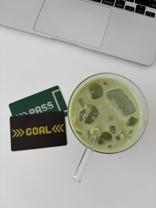 An overhead shot of a glass of iced green tea, a laptop edge, and two pixel-style cards reading 'COASSI' and 'GOAL' on a white desk.