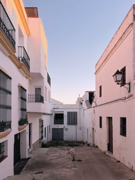 A narrow street lined with white-washed Mediterranean-style buildings, featuring small balconies with wrought iron railings and a traditional wall-mounted lantern under a clear sky.