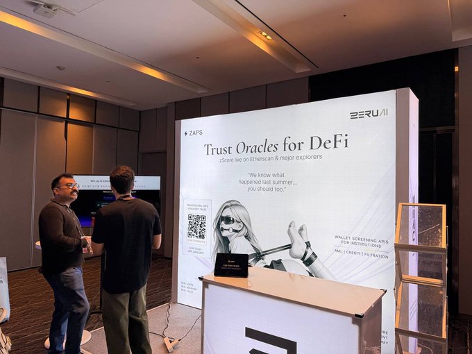 Two men converse at an indoor event booth displaying 'ZERU AI' branding with a poster reading 'Trust Oracles for DeFi zScore live on Ethoscan & major explorers' and 'WALLET SCREENING API FOR INSTITUTIONS'.