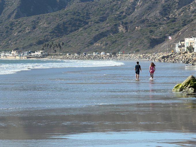 Two people walk along the wet shoreline of a beach with mountains and coastal buildings in the background.