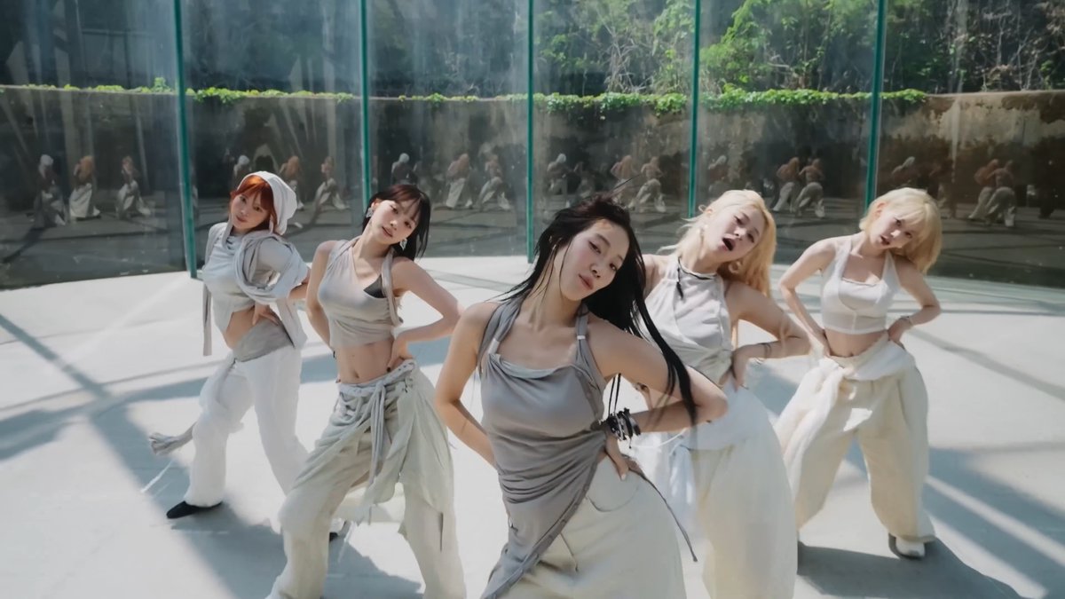 Five young women in coordinated neutral-toned outfits performing a choreographed dance routine in a modern room with glass walls.