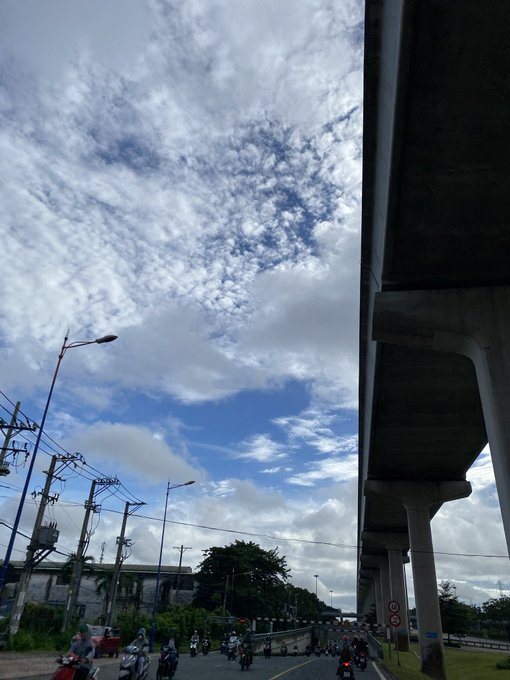 A view looking up at a concrete elevated highway structure on the right and a street lamp on the left, with motorbikes traveling on the road below under a sky filled with scattered clouds.