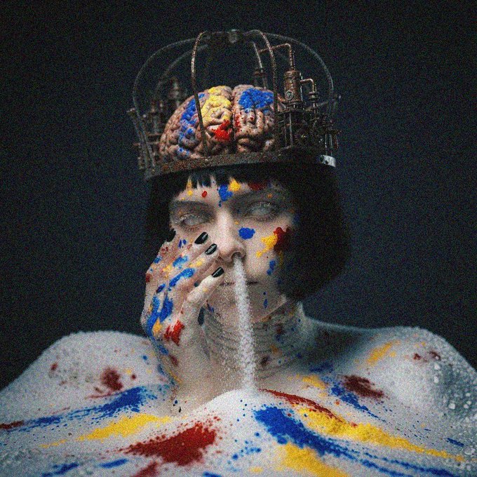 A conceptual portrait of a pale woman with dark bobbed hair, her face and white clothing covered in colorful paint splatters, wearing a metallic headpiece that reveals an exposed brain and spraying a white substance from her mouth.