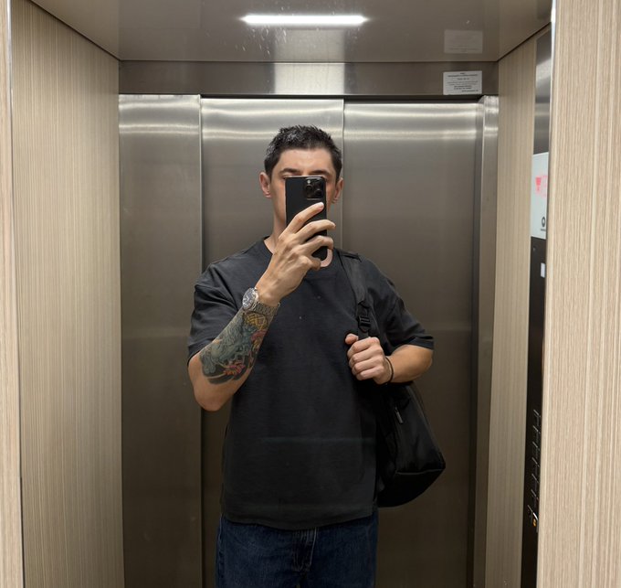 A mirror selfie of a man in a black t-shirt holding a smartphone in an elevator, showing tattoos on his arm.