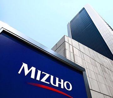 A low-angle shot shows the Mizuho (blue and red) corporate sign in the foreground, with a tall skyscraper behind it under a clear blue sky. The image illustrates the Japanese investment bank referenced in the post, which has adjusted its price target on Strategy ($MSTR).