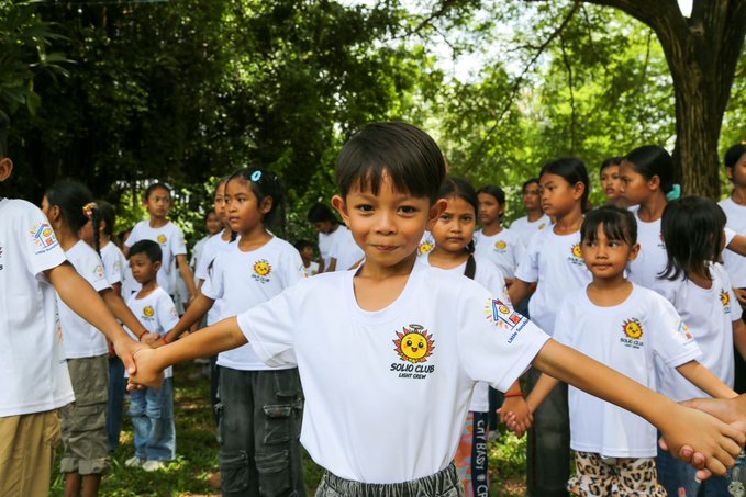 A young boy in a white 'Solar Club' t-shirt smiles at the camera while holding hands with other children in matching shirts, forming a circle outdoors.