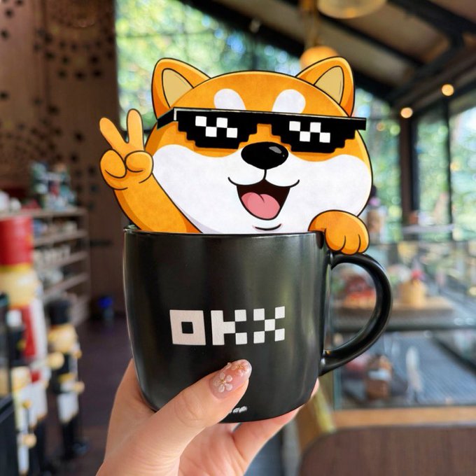 A hand holding a black mug with the 'OKX' logo, featuring a cartoon Shiba Inu dog wearing pixel sunglasses and making a peace sign popping out of the cup.