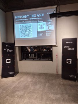 Photo of a pop-up event booth titled 'INTO ORBIT' featuring a projected QR code slide flanked by black vertical banners with QR codes and red arrow accents.