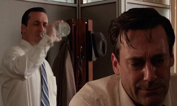 Two men in a 1960s office setting, one drinking from a glass in the background while the other, Don Draper from Mad Men, sits in the foreground looking somber.