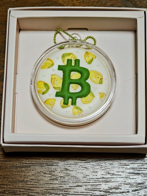 A round Bitcoin-themed ornament with a green "B" logo and yellow lemon patterns, displayed in its packaging box with a green hanging cord.