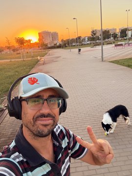 A selfie of a smiling man wearing a Huawei cap, glasses, and headphones in an urban park at sunset, pointing his finger toward a black and white dog walking on the paved path behind him.