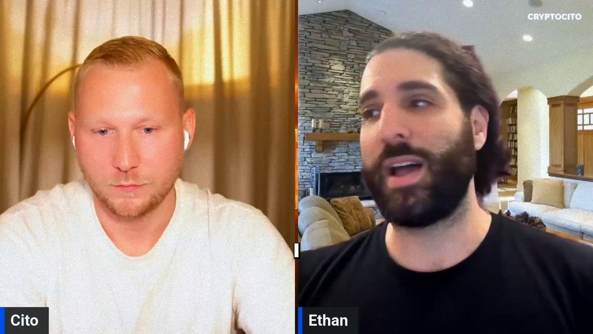 The image shows a man wearing a white shirt having a video call with another person, presumably Ethan Buchman, who is also wearing a black shirt. They are both looking at each other through their computer screens as they engage in conversation. The background of the image reveals that it's taking place in someone's living room or bedroom area.