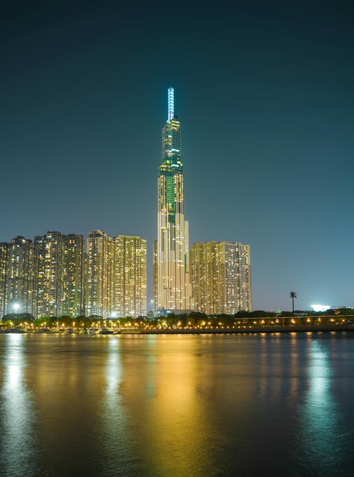 The image features a tall building with lights shining brightly against the dark sky, creating an impressive skyline at night. The cityscape includes several other buildings of varying heights, contributing to the overall grandeur of the scene. In the foreground, there is water that reflects the lights from the skyscraper and the surrounding area, adding depth and dimension to the image. The post contains a message encouraging people to "Have a good night all Dream will come true w @quipnetwork @wallchain g9".