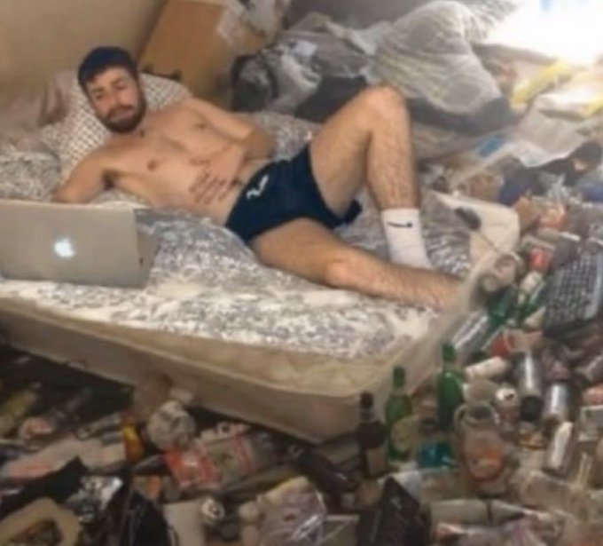 A shirtless man in dark athletic shorts and white Nike socks lying on a mattress in an extremely cluttered room filled with trash, bottles, and cans, with an open Apple laptop next to him.