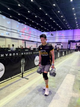 A man wearing a black t-shirt with 'LETHAL' on it and carrying dumbbells walks through a large convention center with 'HYROX' banners lining the floor.