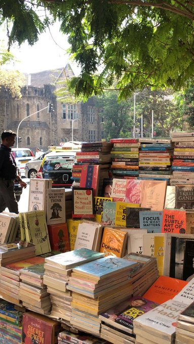 The image shows an outdoor book sale with a large stack of books displayed prominently. The books are arranged in various sizes and colors, creating an inviting atmosphere for potential buyers. A person can be seen walking past the book sale, adding life to the scene. In the background, there is a car parked nearby, indicating that this event might be taking place in a busy area or near a parking lot. The overall setting suggests that the book sale could be part of an initiative by @CNPYNetwork to promote reading and learning among its members while fostering connections within the community.