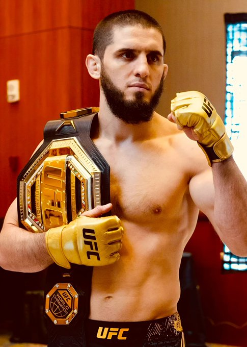 Islam Makhachev poses with his UFC championship belt, wearing gold UFC gloves, in what appears to be a post-fight or pre-fight moment. The image connects to the post as Makhachev is the fighter in the high-stakes $293,000 bet on Polymarket predicting his victory.