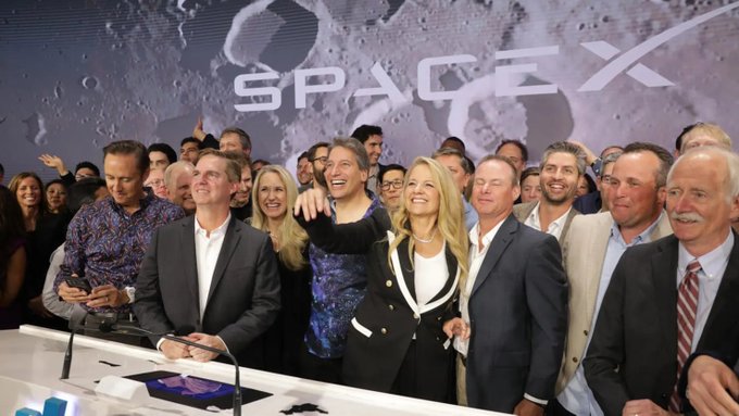 The image shows a group of people gathered around a table at a conference or event. They appear to be posing for a photo with their hands raised towards the sky as if they are pointing at something interesting, possibly related to the SpaceX company. The background features a large screen displaying the SpaceX logo and the words "Passive" and "$SPCX $BTC". This suggests that the attendees might be discussing or learning about the latest developments in space exploration or investing in the company's stock.