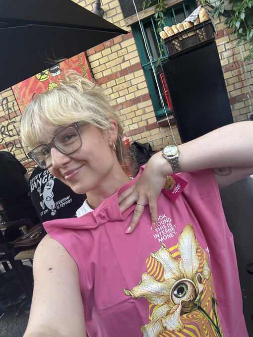 A blonde woman wearing glasses smiles for a selfie, modeling a pink sleeveless shirt featuring a large eye graphic and the text 'LOOK AROUND. THIS IS INTERNET MONEY'.