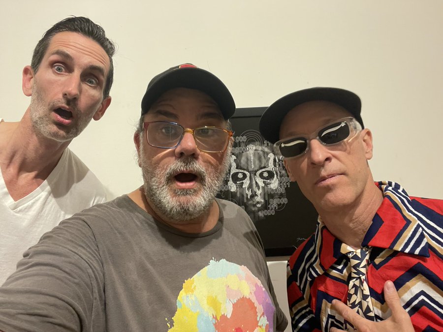 Three men pose for a selfie in front of a wall-mounted skull artwork; one wears a white shirt with a surprised expression, the middle man has a beard and cap looking shocked, and the third wears sunglasses and a colorful striped shirt.