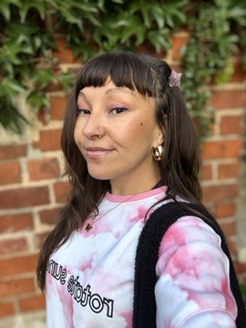 A portrait of a young woman with dark hair and bangs, wearing a pink and white patterned shirt and a backpack, smiling slightly with a brick wall and green leaves behind her.