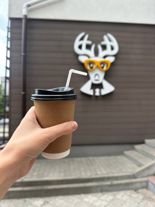 A hand holds a brown paper coffee cup with a black lid and white straw in front of a building featuring a deer logo wearing yellow sunglasses.