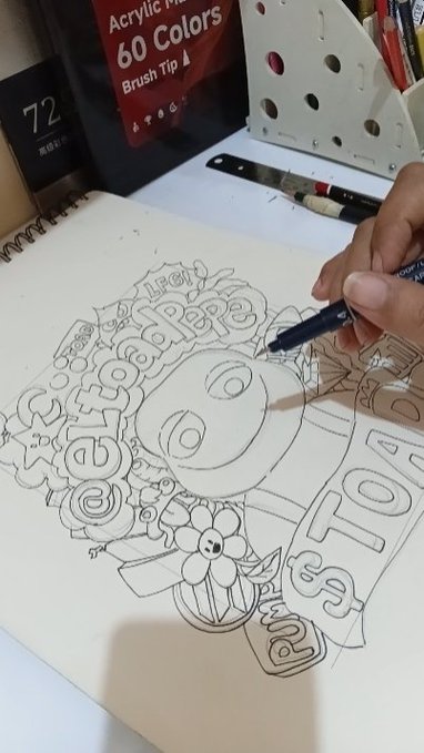 A hand sketches a detailed black-and-white drawing of a character with text bubbles reading 'Leg', 'Feedipe', 'eat', 'TO', 'ame', alongside flowers and hearts, with a 60-color acrylic paint set visible in the background.