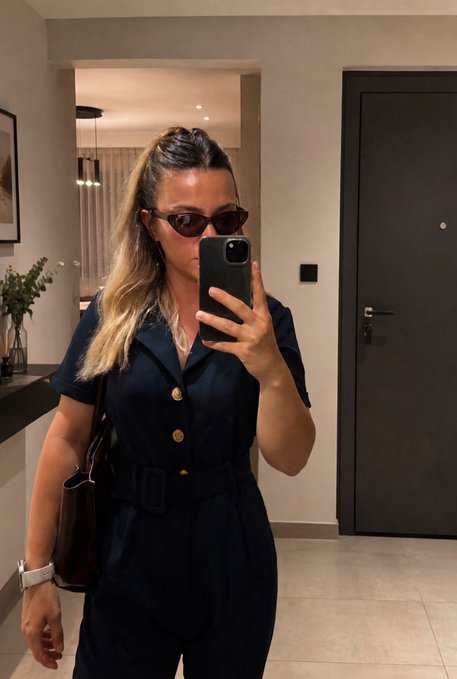A woman wearing a dark navy belted jumpsuit and sunglasses takes a mirror selfie with a smartphone in a modern interior.