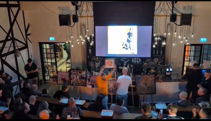 The image shows an event taking place at a conference, where people are gathered around a large screen displaying artwork for sale. A group of artists can be seen standing near the screen, possibly discussing or showcasing their work. The audience consists of several individuals who have come together to appreciate and participate in this unique auction experience.