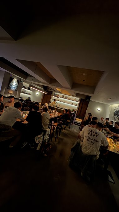 A dimly lit bar or restaurant interior with patrons seated at the counter and tables, where one person wears a white t-shirt reading 'SAINT BABE' on the back.
