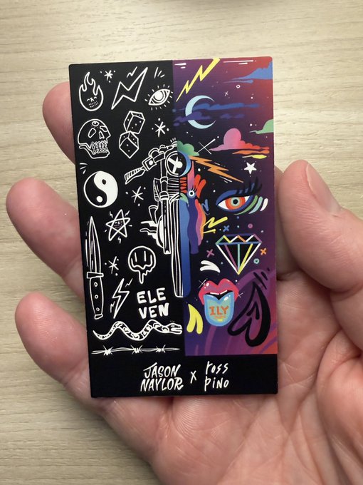 A hand holding a small artist trading card titled 'ELEVEN' by Jaron Naylor and Ross Pino, featuring illustrations of a knife, skull, yin-yang, and other mystical symbols.