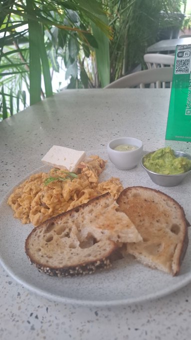 A hearty breakfast spread featuring scrambled eggs, a block of feta cheese, slices of toasted sourdough bread, and small bowls of butter and guacamole sits on a speckled table, paired with a green menu displaying a QR code and a "WiFi" sticker. The image complements the post's relaxed Monday morning vibe of enjoying a meal while catching up on CT news.