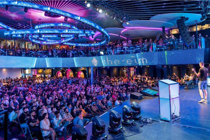 A large crowd gathered inside a multi-level venue called "The e°um" with "ethereum" branding displayed on circular overhead screens, while a speaker presents on stage under purple and blue lighting.