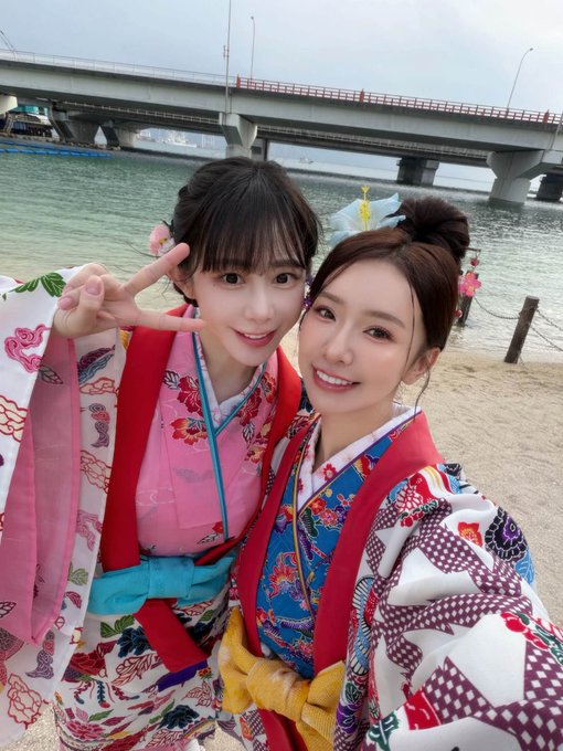 Two smiling women in colorful traditional kimono take a selfie outdoors, with a body of water and a bridge visible in the background.