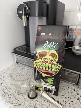 A BAK vending machine setup on a kitchen counter next to a Keurig coffee maker, with a clear cup and a coffee pod placed in front of it.