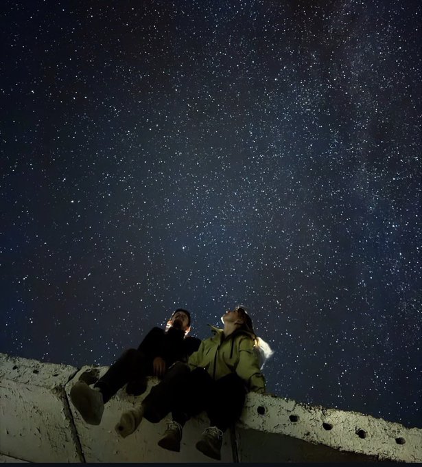Two people sit on a concrete ledge at night, looking up at a vast starry sky, with one person illuminated by a phone screen.