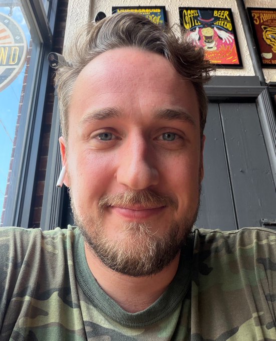 A selfie of a smiling man with light brown hair, a beard, and an earring, wearing a camouflage t-shirt, posed in front of colorful bar signs advertising drinks like "Coffee City Beer Checked" and "Panther." The casual, upbeat vibe matches the cheerful "gm grinders happy hump day!" morning greeting.